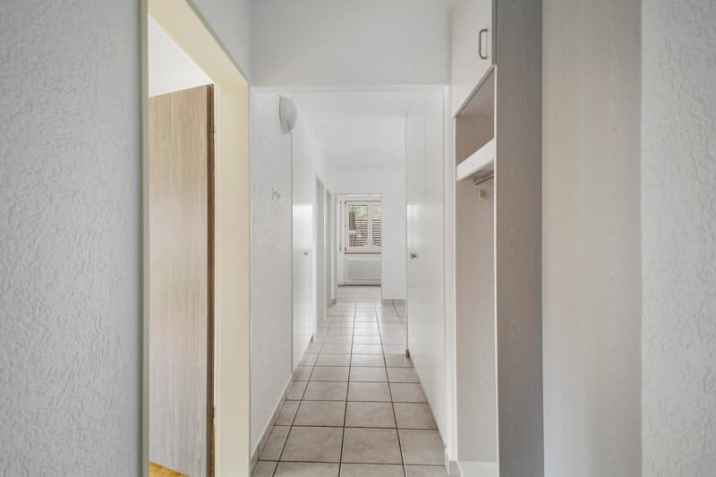 The image shows a long, narrow hallway with white walls and a tiled floor. The hallway has several doors leading to other rooms, and a window at the end providing natural light. The hallway appears to be part of a larger residential or commercial space.