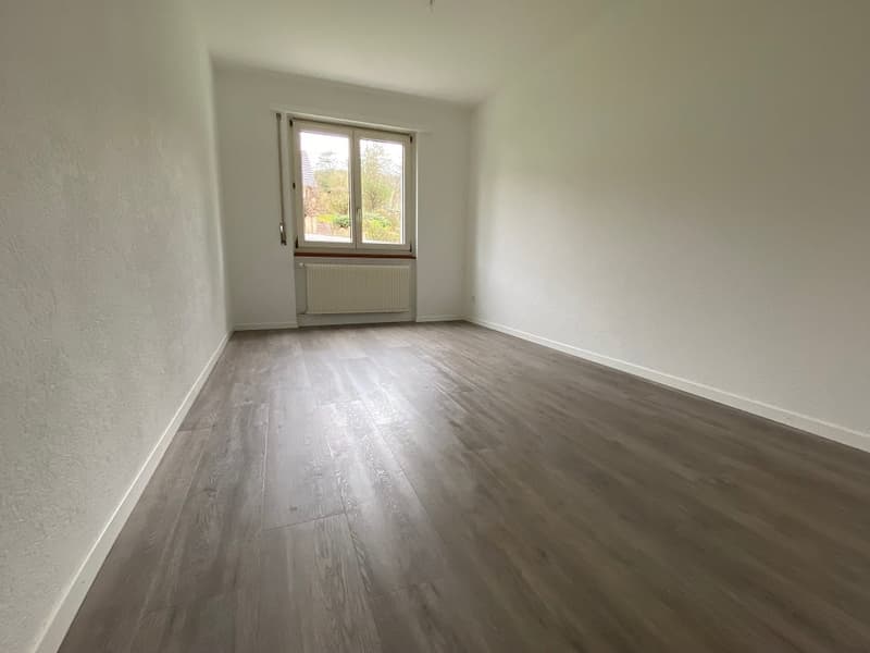 Spacious bedroom with white walls, hardwood flooring, and a large window overlooking the outdoor area.