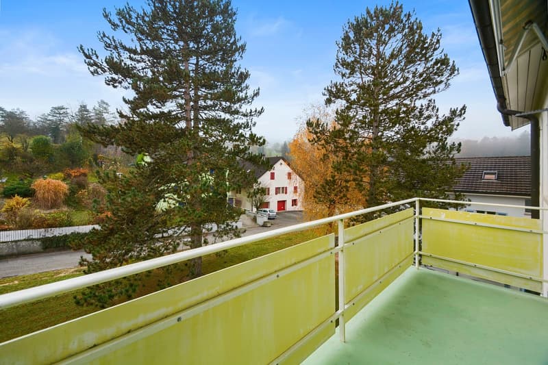 The image shows a balcony or terrace overlooking a scenic autumn landscape. The balcony has a yellow railing and offers a view of a residential building with a red roof, surrounded by pine trees and other foliage in various fall colors.
