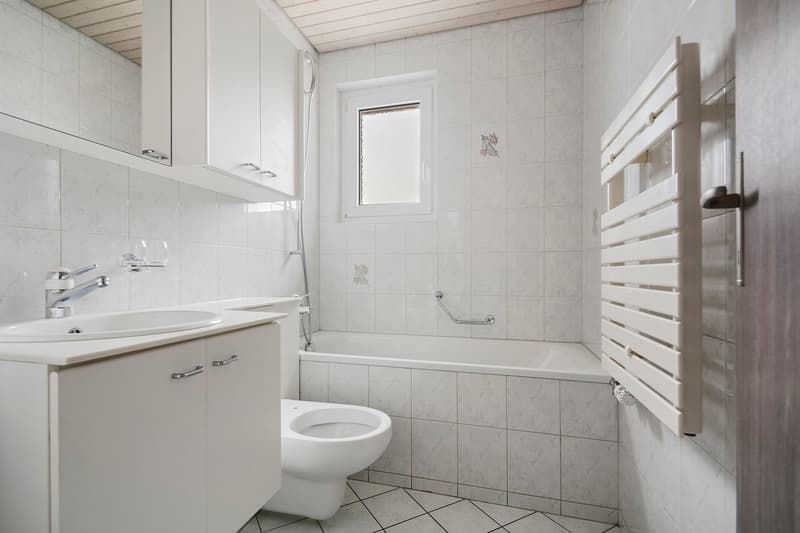 Tiled bathroom with a vanity cabinet, sink, toilet, and a bathtub with a shower curtain. The room has a window and a towel rack on the wall.