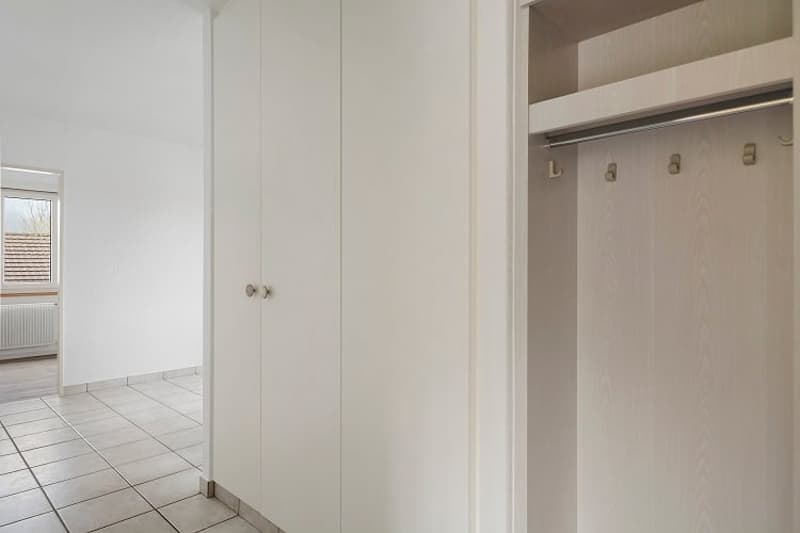 The image shows a hallway with white walls, a tiled floor, and built-in closets or cabinets along one wall. The hallway appears to be part of a larger living space, with an open doorway visible in the background.