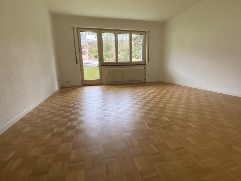 Spacious living room with hardwood parquet flooring, large windows providing natural light, and a radiator along the wall.