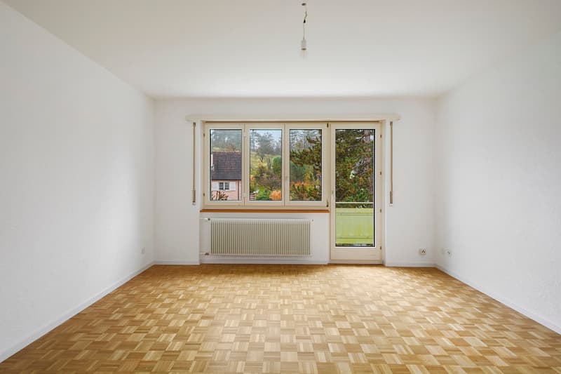 Spacious living room with large windows overlooking a garden, hardwood parquet flooring, and a radiator on the wall.