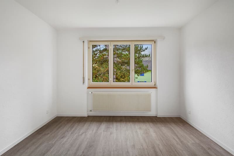 Spacious bedroom with large windows overlooking a wooded area, hardwood-style flooring, and a radiator heater on the wall.