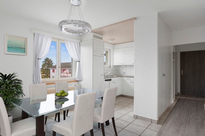 white walls, white ceiling, glass table, 6 chairs, modern chandelier, white kitchen, white radiator, window with white curtains, picture frame, wooden floor
