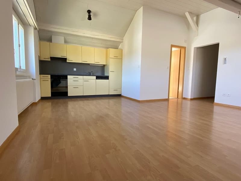 Empty room with wooden floor, white walls, kitchen with cabinets, sink, and dishwasher, windows, and a door.
