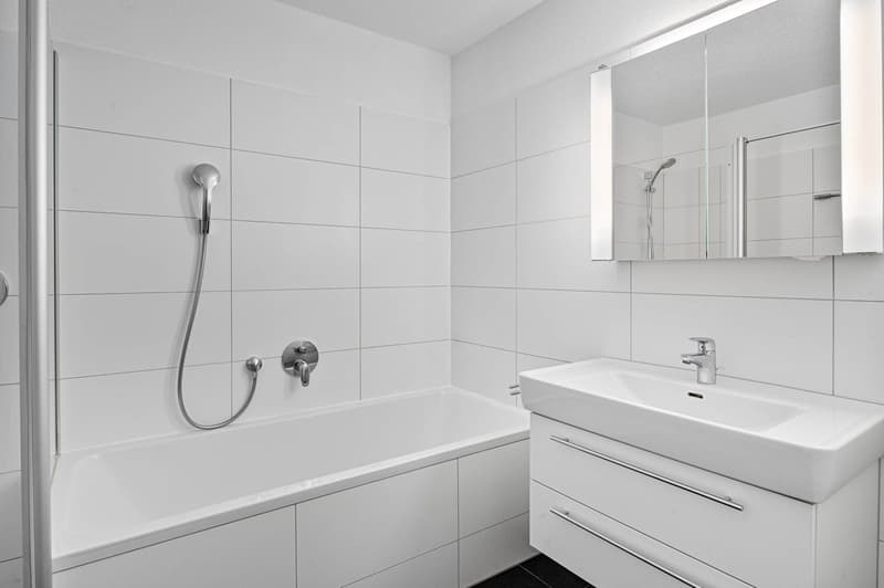 White tiled walls, white vanity, white bathtub, white sink, mirror, showerhead, cabinet