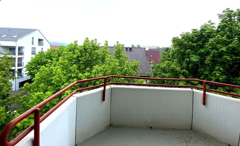 Balcony with red railings, concrete floor, view of trees and buildings