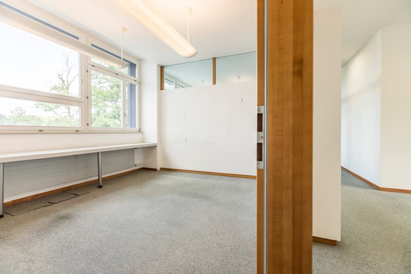 This image shows a spacious room with large windows, providing ample natural light. The room has a carpeted floor and white walls, with wooden accents and trim. There is a built-in desk or workstation along one wall, and the room appears to be set up as a