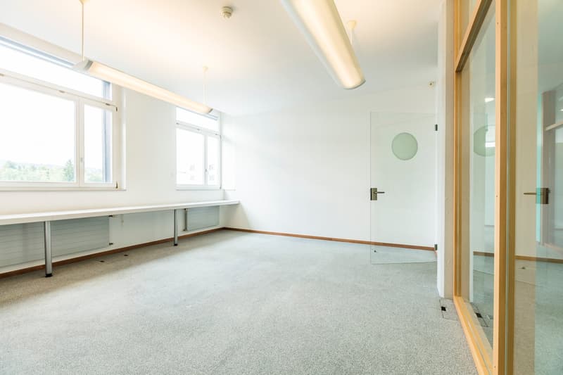 Large open room with white walls, carpeted floor, and built-in desk along the window. The room has overhead lighting and a glass door leading to another space.