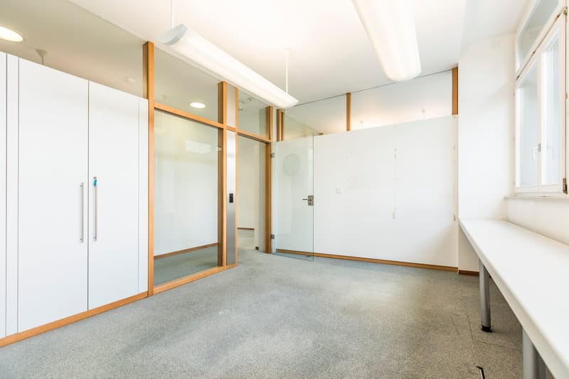 The image shows a spacious office space with white walls, a gray carpeted floor, and large glass windows and doors. The room has a minimalist design with wooden accents on the frames of the glass partitions. There is a small window on the side wall and a
