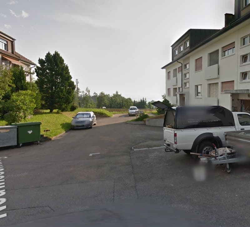 The image shows a residential area with apartment buildings, trees, and a paved parking area with cars parked. There appears to be a trailer or utility vehicle parked in the lot as well.