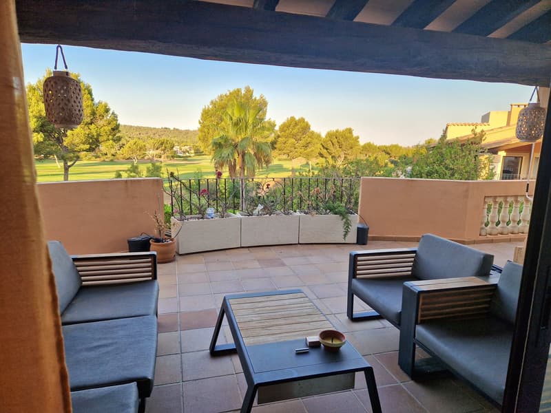 covered terrace, large couch, two chairs, coffee table, two hanging lamps, garden view, golf course in the distance