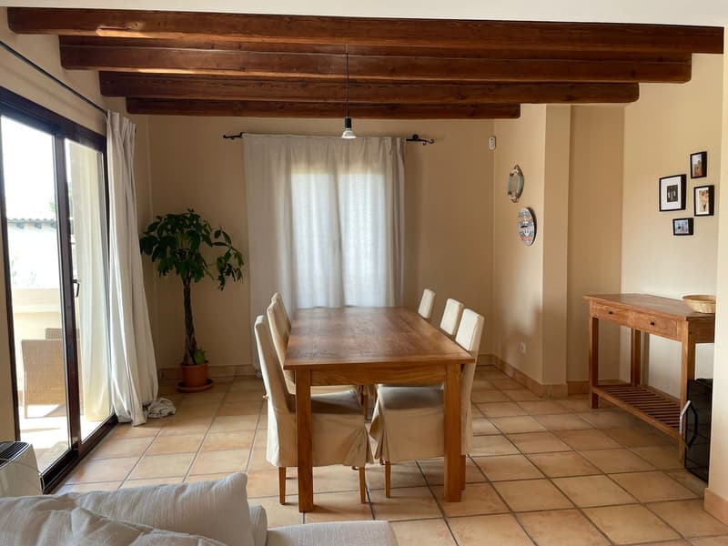 Dining room with wooden table, white chairs, beige walls, and wooden beams on the ceiling. It also features glass doors, a potted plant, a white sofa, and a side table with a basket on top.