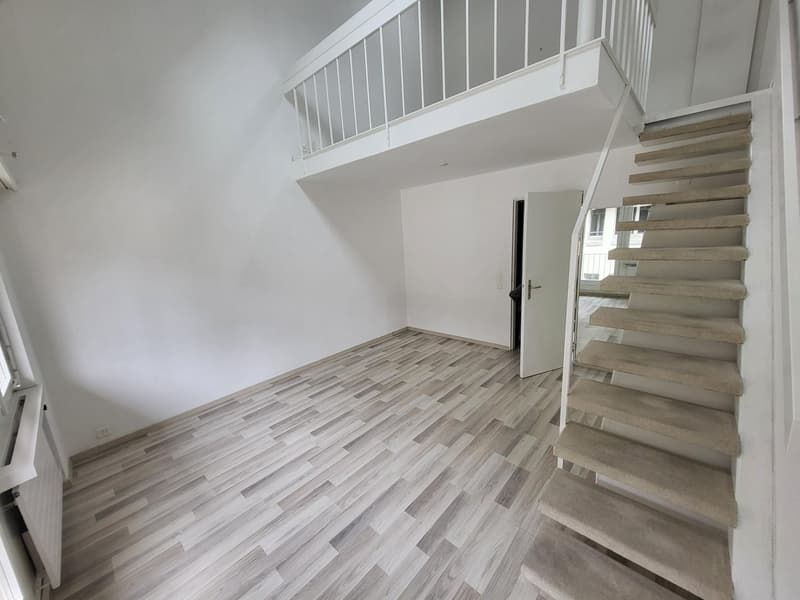 Empty room, white walls, gray tiled floor, wooden stairs, closed door, white radiator, metal balcony railings