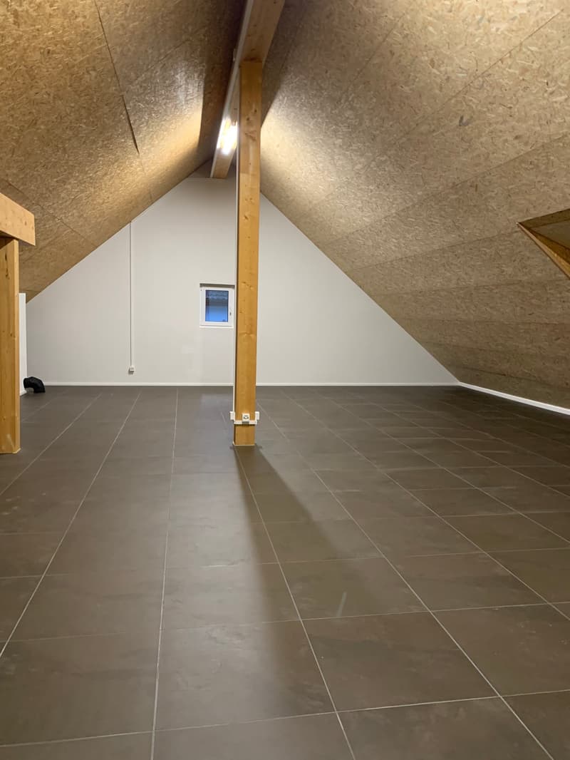 The image shows an attic space with a slanted ceiling and wooden beams. The floor is covered with gray tile, and there is a small window visible. The space appears to be unfinished, with exposed insulation and structural elements.