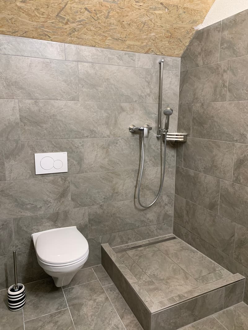 The image shows a modern bathroom with gray tile walls and floor. It has a white toilet, a shower with a handheld showerhead, and a built-in bench or shelf. The ceiling appears to be made of OSB (oriented strand board) material.