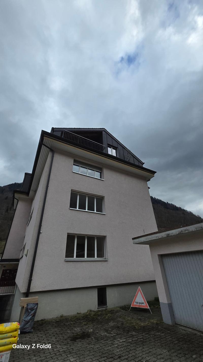 A multi-story apartment building with a gray exterior, large windows, and a balcony visible on the top floor. The building appears to be located in a mountainous area with cloudy skies in the background.