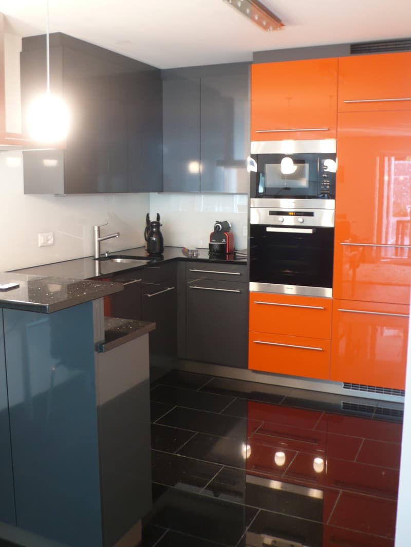 Modern kitchen with orange cabinets, black countertops, stainless steel appliances, under-cabinet lighting, faucet, sink, microwave, oven, black tiled floor