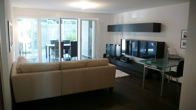 A beige sofa with cushions, an entertainment center with two televisions, glass table, glass doors leading to a balcony with table and chairs