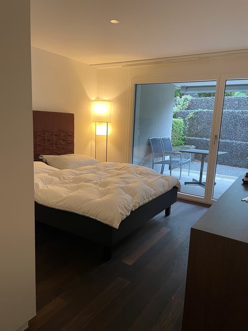 A bedroom with a bed, a lamp, and a desk. The bed has two pillows, a duvet, and a headboard. The lamp is placed on the side of the bed. The desk has a chair and a table. A sliding glass door leads to a balcony with a metal railing, a table, and chairs. Th