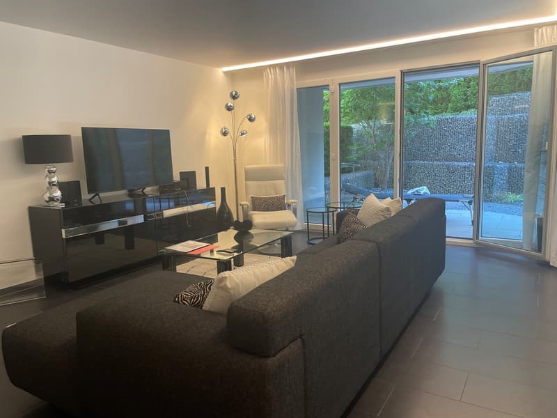 Spacious living room with large windows overlooking a balcony or terrace, dark gray sofa, black entertainment unit with various decor items, and a floor lamp with multiple bulbs
