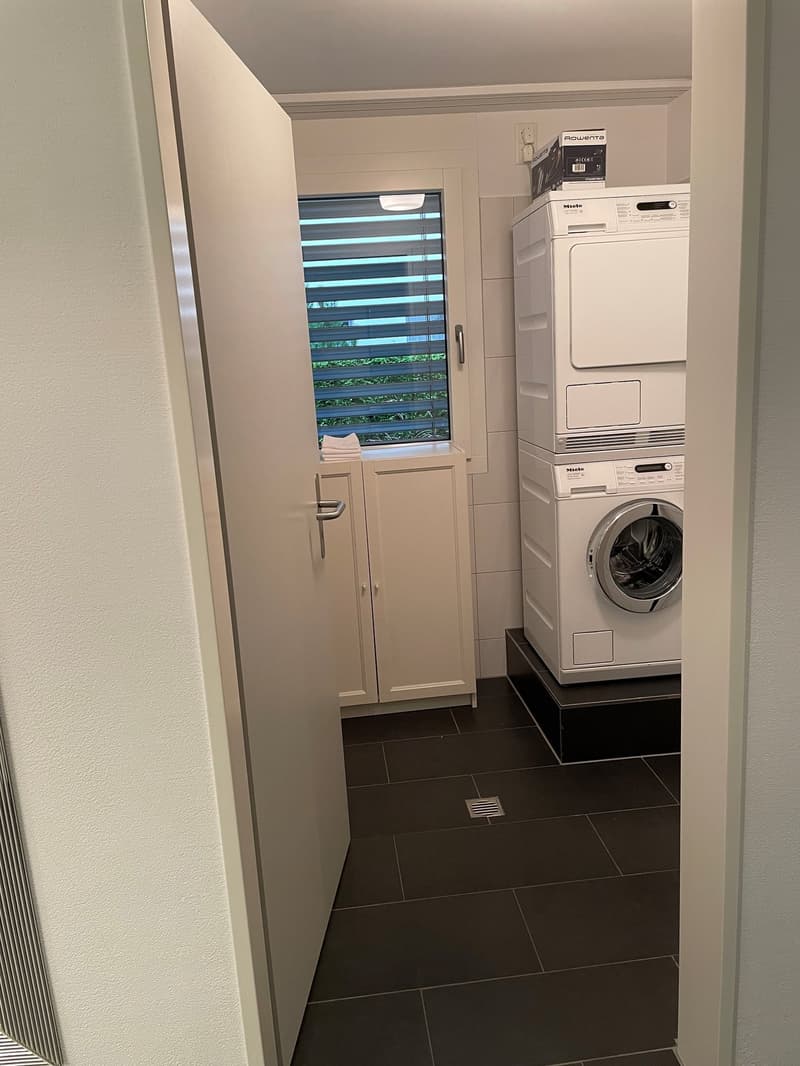 The image shows a utility room with a washing machine and a dishwasher. The room has a tiled floor and a window with blinds. There are also some storage cabinets in the room.