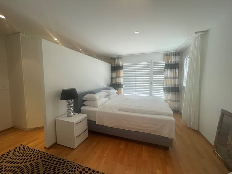 Spacious bedroom with hardwood flooring, white walls, large windows with blinds, a king-size bed, nightstands, and a decorative lamp.