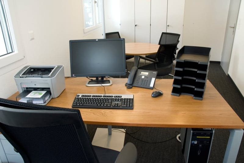 A desk with a computer, keyboard, mouse, printer, telephone, and other office equipment. There are chairs and a round table in the background.