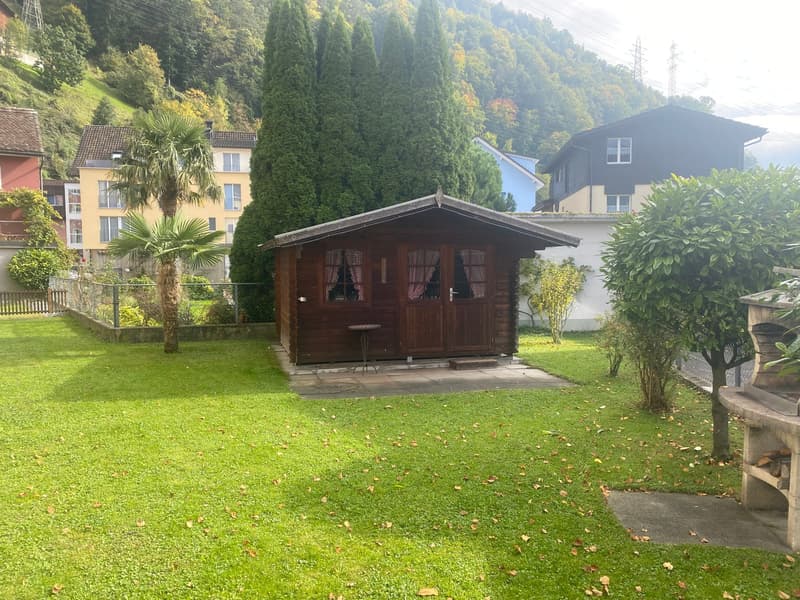 Wooden house on a grassy yard, surrounded by other buildings and trees, with a table in front of it and a grill in the background.