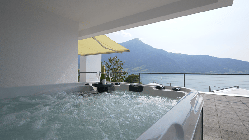 Outdoor hot tub on balcony, water running, clear water, metal railing, yellow awning, view of lake and mountains, wine bottle and two glasses on table