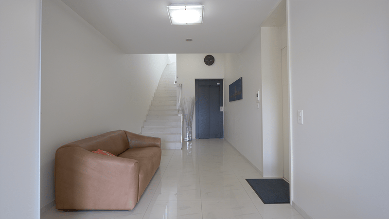 White walls, white tiled floor, staircase, blue entrance door, mounted clock, decorative piece, artwork on wall, brown leather sofa with pillow, modern minimalist design
