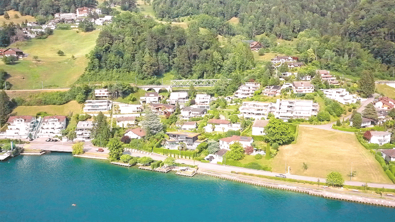 cluster of residential buildings, some with balconies, set around a lake, greenery, hillsides, bridge in distance