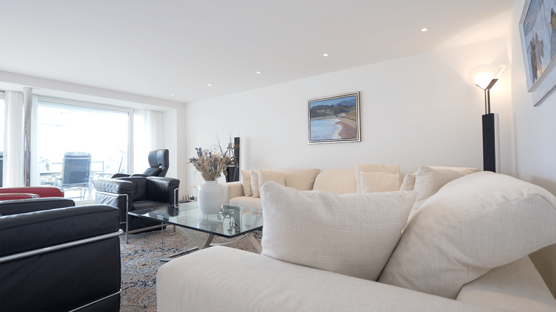 Modern living room with white walls and modern decor, glass center table, multiple white sofas, glass windows, and framed pictures