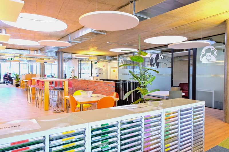 Modern open office with white tables, round chairs, circular lights, a brick wall, plants, and a metal barrier. Various office supplies are organized in a row of drawers.
