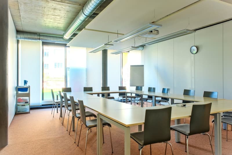 large room with conference tables and chairs, whiteboard, ceiling lights, clock, windows