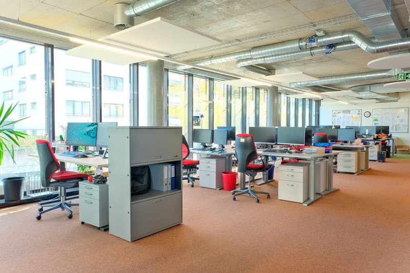 Modern office room, desks, computer monitors, chairs, filing cabinets, red office chairs, trash bins, potted plants