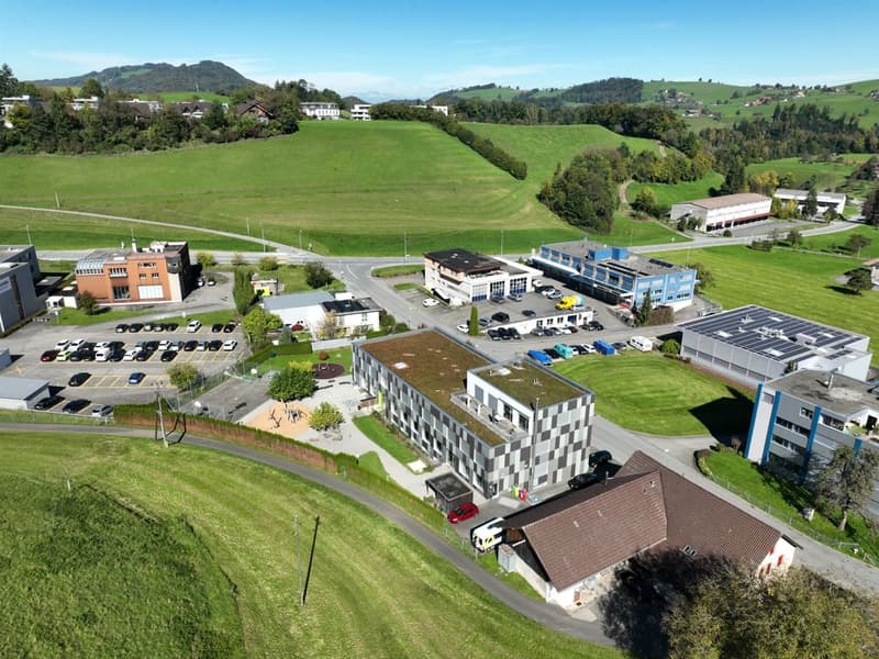 Complex of buildings in an urban area, multiple parking spaces, grassy fields, and trees.