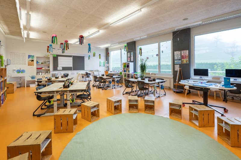 Empty classroom with orange floor, various tables and chairs, a round carpet, a plant, and monitors on desks.