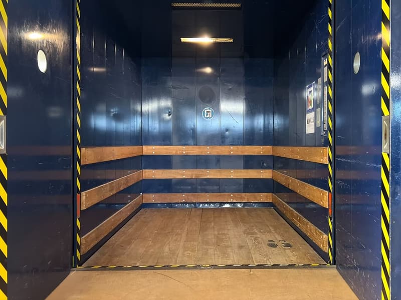 Empty elevator, wooden flooring, blue walls, wooden benches, circular markings, and safety features