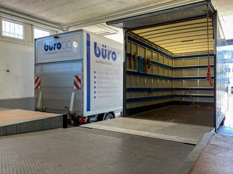 An open moving truck is parked in a garage. There is a ramp attached to the truck and the interior is shown with shelving and equipment.