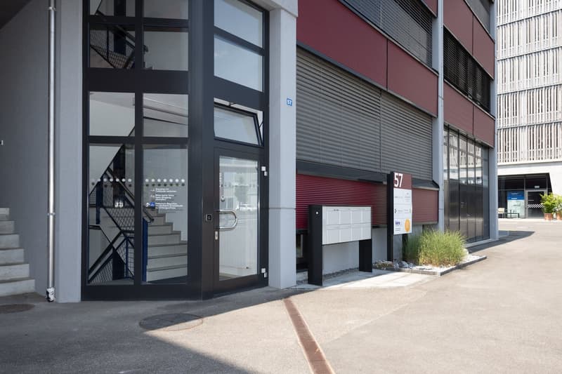A modern building entrance with black glass doors, metal railings on staircases, mailbox, a ground-level door, and a parking area