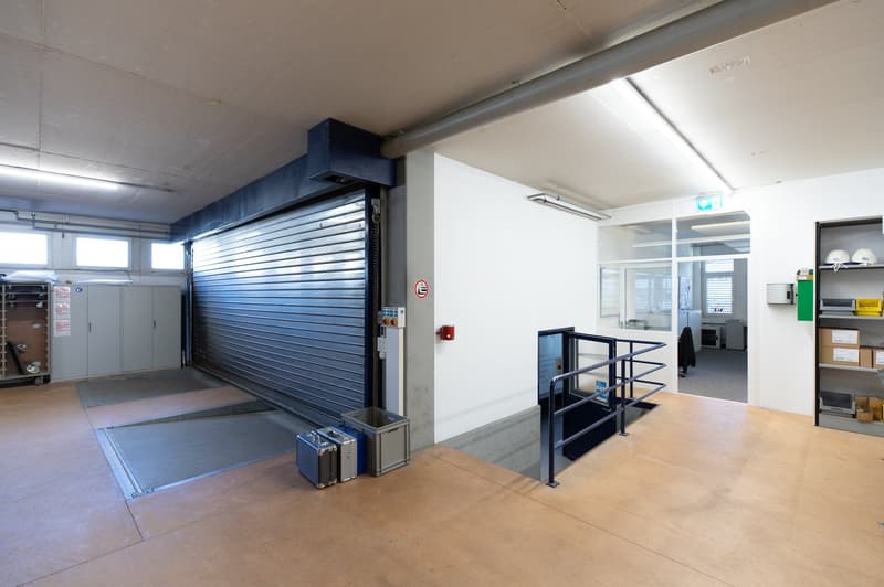 Large open area, roll-up door, concrete flooring, metal railings, closed off space, storage boxes, office equipment, signboards
