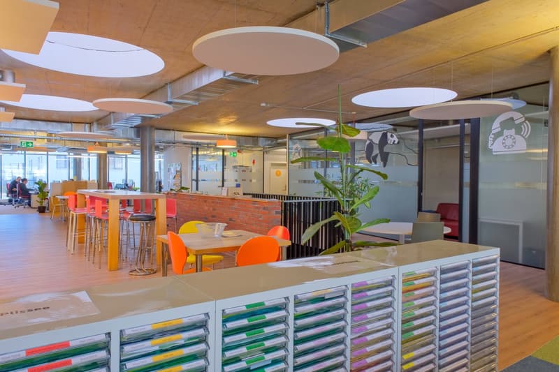 Open area with desks, chairs, and plants. Features large round ceiling lights and a partition with a brick wall.