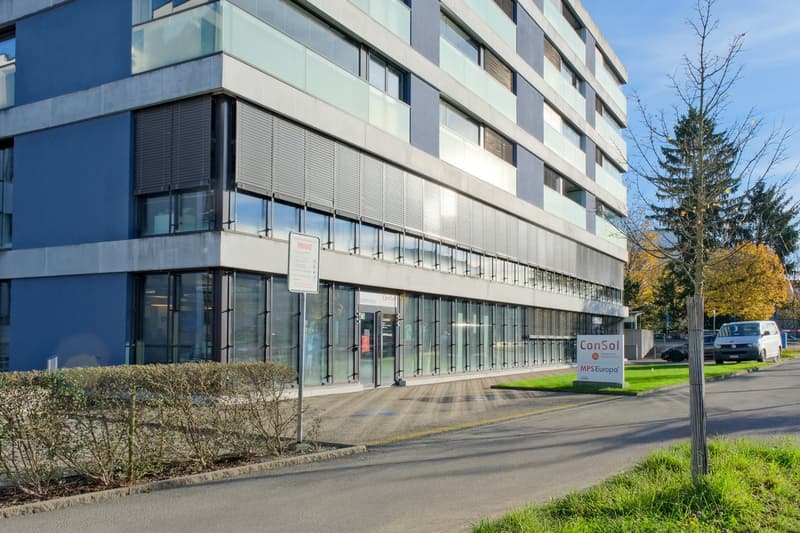 4 story office building with blue facade, glass windows, glass entrance door, shrubs in front, cars parked on the side