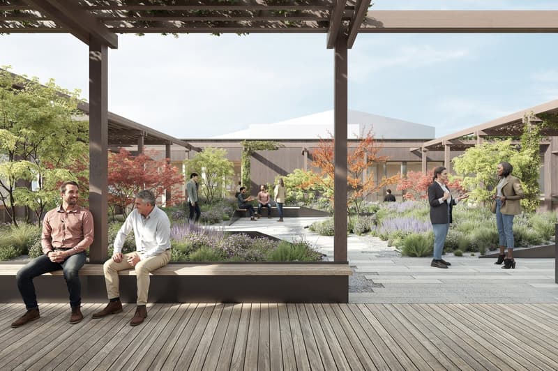 The image shows an outdoor public space with a wooden deck, covered by a pergola structure. There are several people sitting on the deck, and others walking around the landscaped garden area with trees, shrubs, and flowers. The overall design appears to b