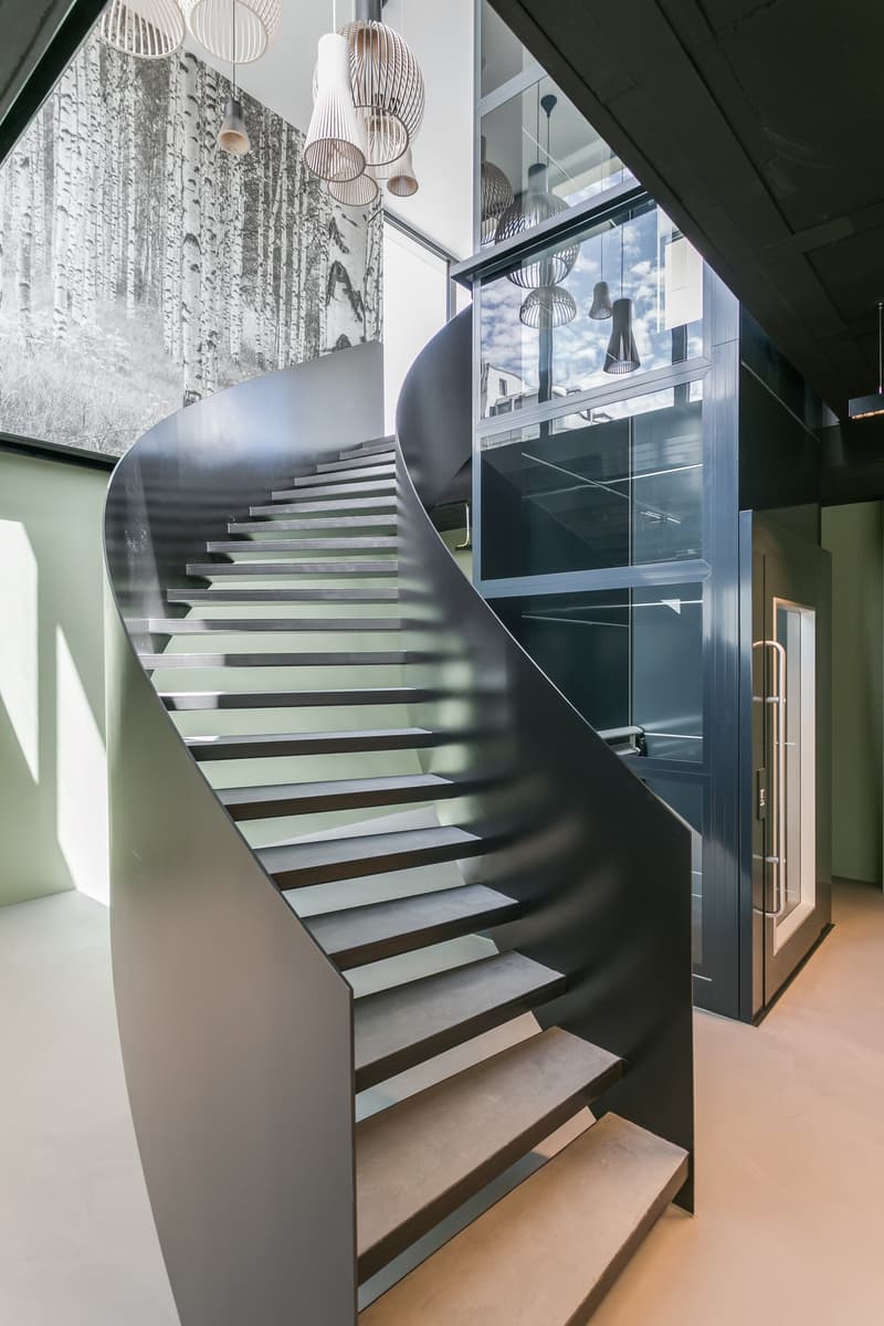Modern and stylish interior with a curved staircase made of metal and wood, suspended lighting fixtures, and large windows providing a view of the exterior.