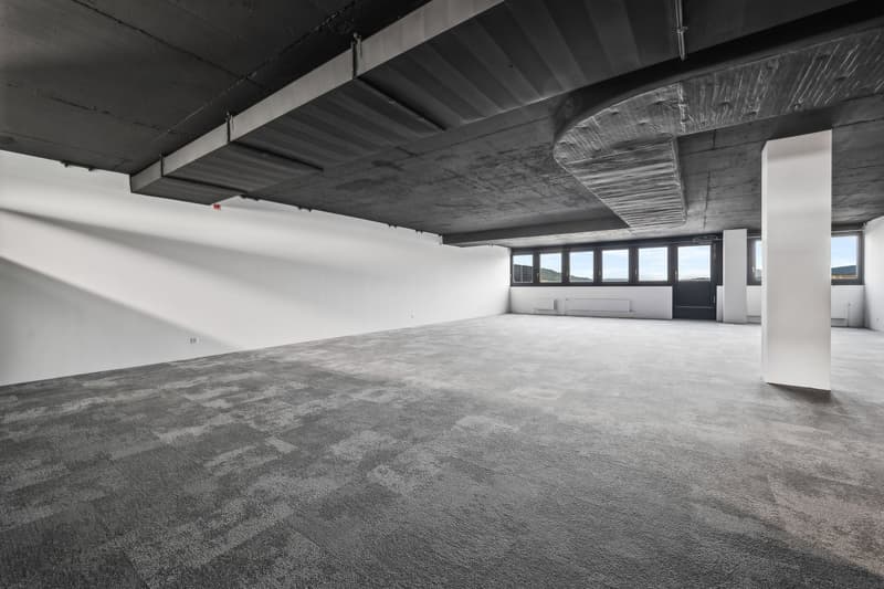 Large open-plan office space with exposed concrete ceiling and floor, white walls, and large windows providing natural light. The space appears to be unfinished or under renovation, with no furniture or equipment visible.