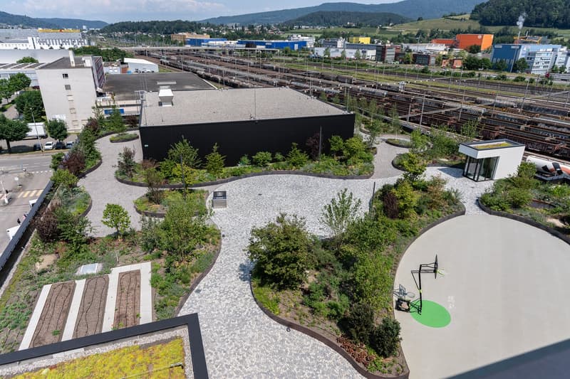 This image shows a large outdoor public space with a paved walkway, landscaped gardens, and various seating areas. The space appears to be located near a train station or industrial area, with railway tracks visible in the background. The design features