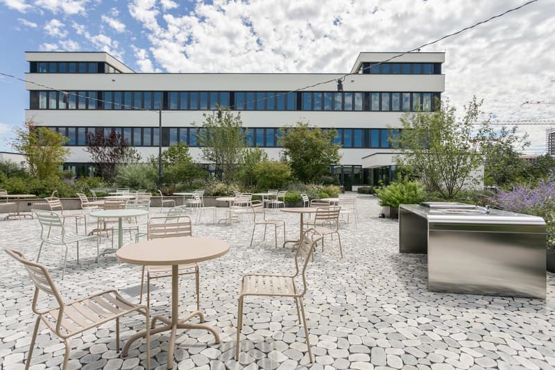 Modern multi-story office building with large windows, surrounded by an outdoor seating area with tables and chairs on a paved stone surface. The building has a clean, minimalist design with a mix of glass and concrete elements. Trees and landscaping are